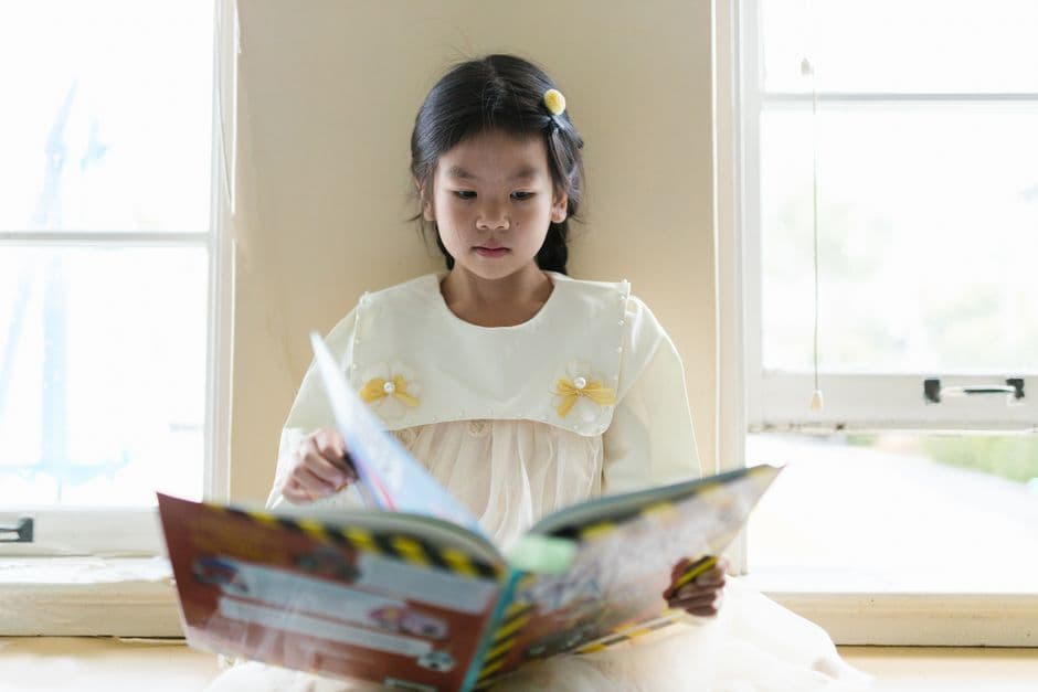 A primary-school child reading a book by a sunlit window, relaxed and absorbed in the page.