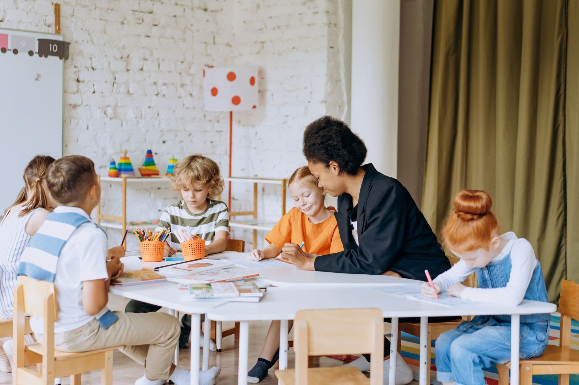 An educator working with a small group of primary-school children at a shared table with books and pens.