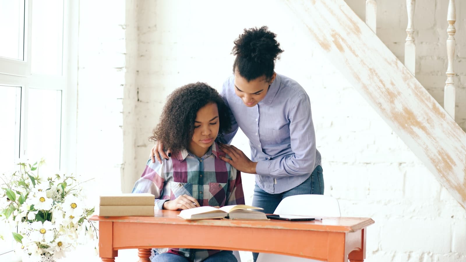 A parent standing with a reassuring hand on her child’s shoulder as the child works at a desk by a bright window.